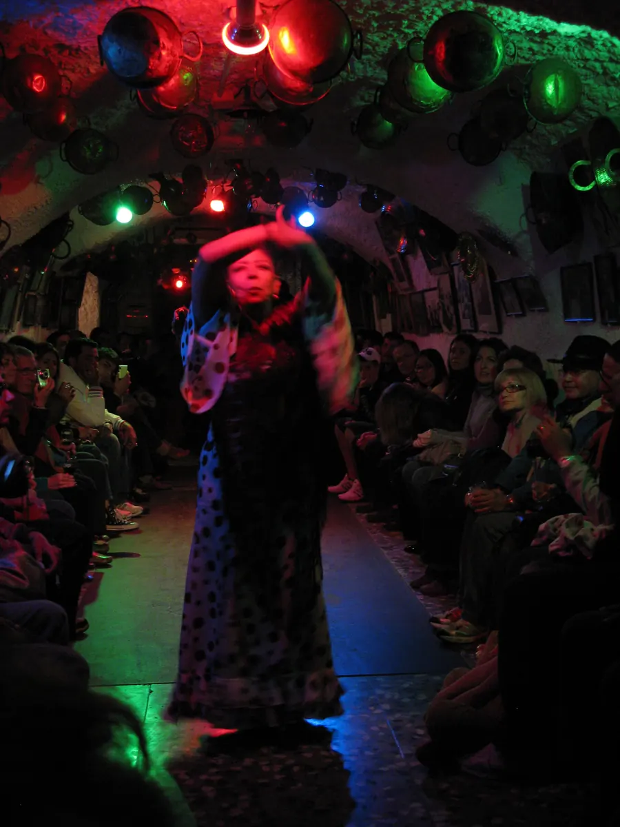 A dancer in a long polka-dot dress with raised arms, lit in red and green, performing in a whitewashed Sacromonte cave with guests seated along the walls