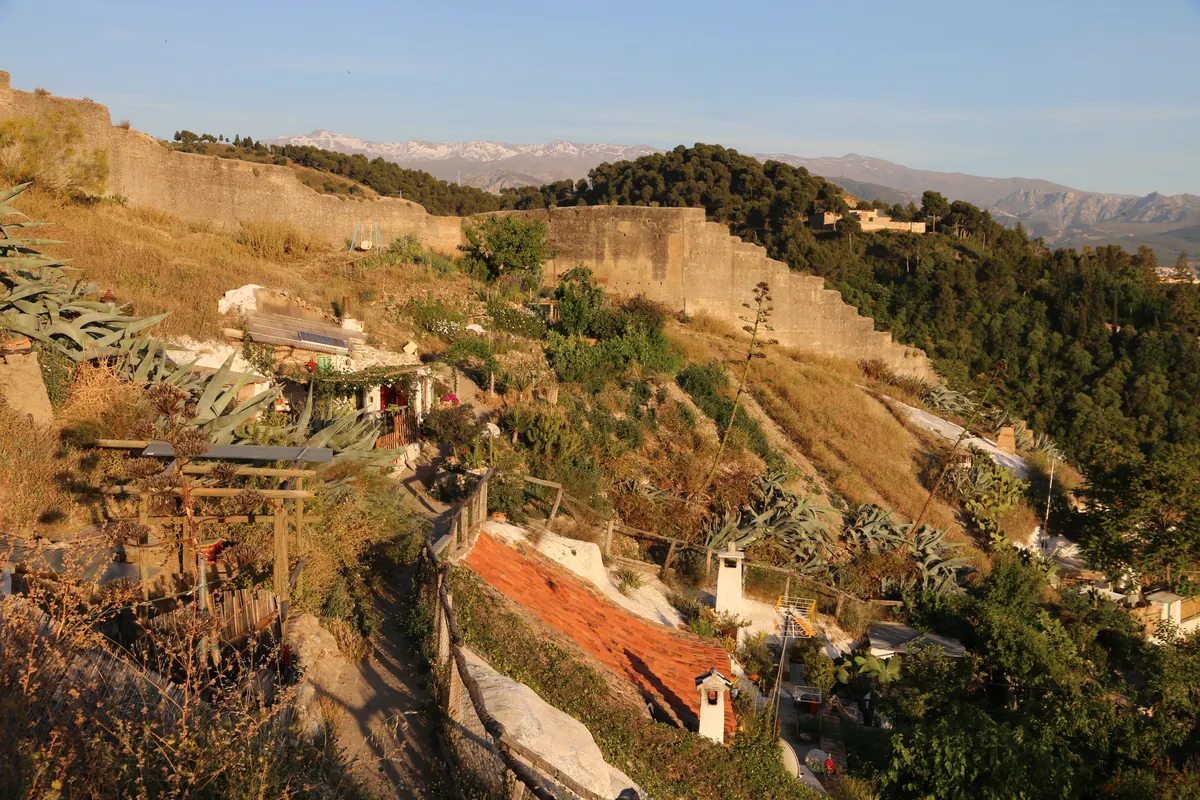 Cave homes and gardens on the Sacromonte slope, with a stone wall above and the Sierra Nevada in the distance