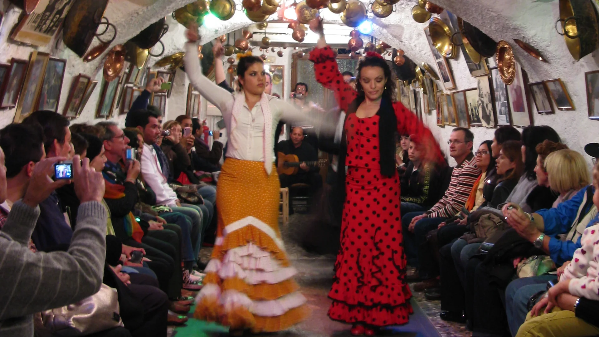 Two flamenco dancers, one in a white blouse and ochre skirt, one in a red polka-dot dress, performing in a whitewashed Sacromonte cave hung with copper pots, with a guitarist behind them and guests seated along the walls
