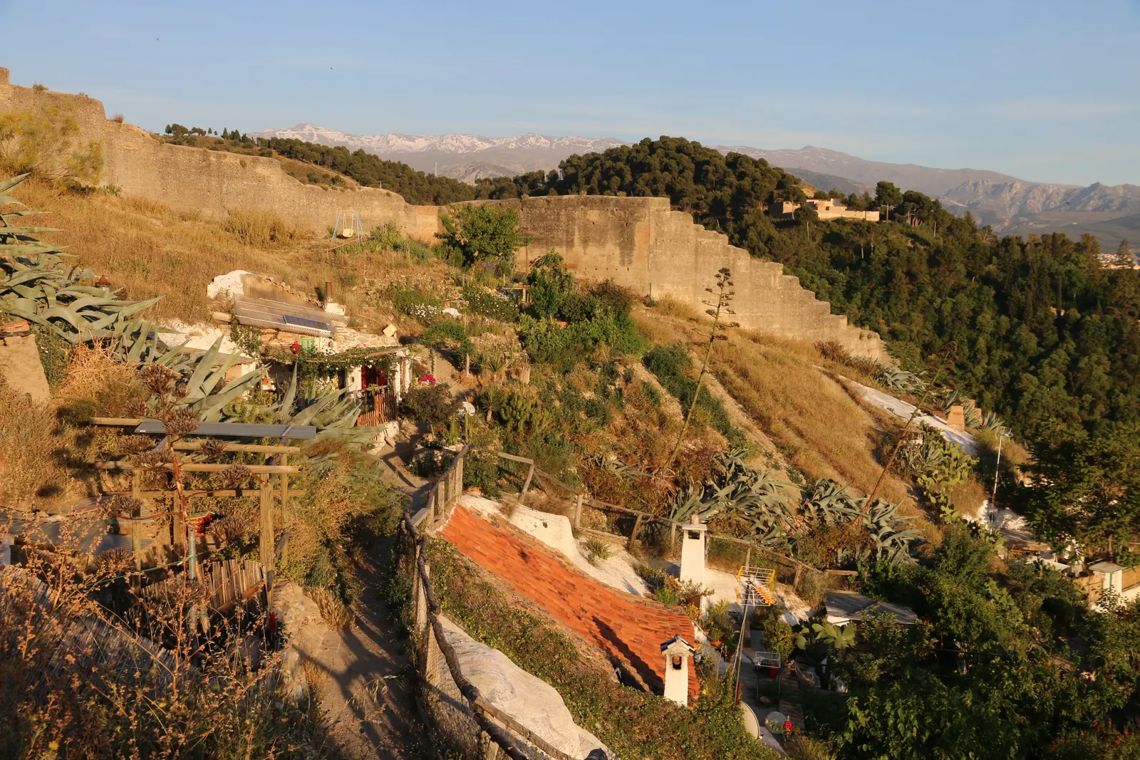 Cave homes and gardens on the Sacromonte slope, with a stone wall above and the Sierra Nevada in the distance